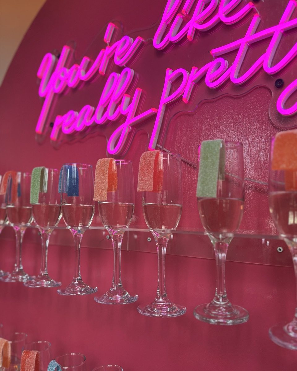 A champagne wall of colorful sugar-rim flutes under a pink neon sign at an event