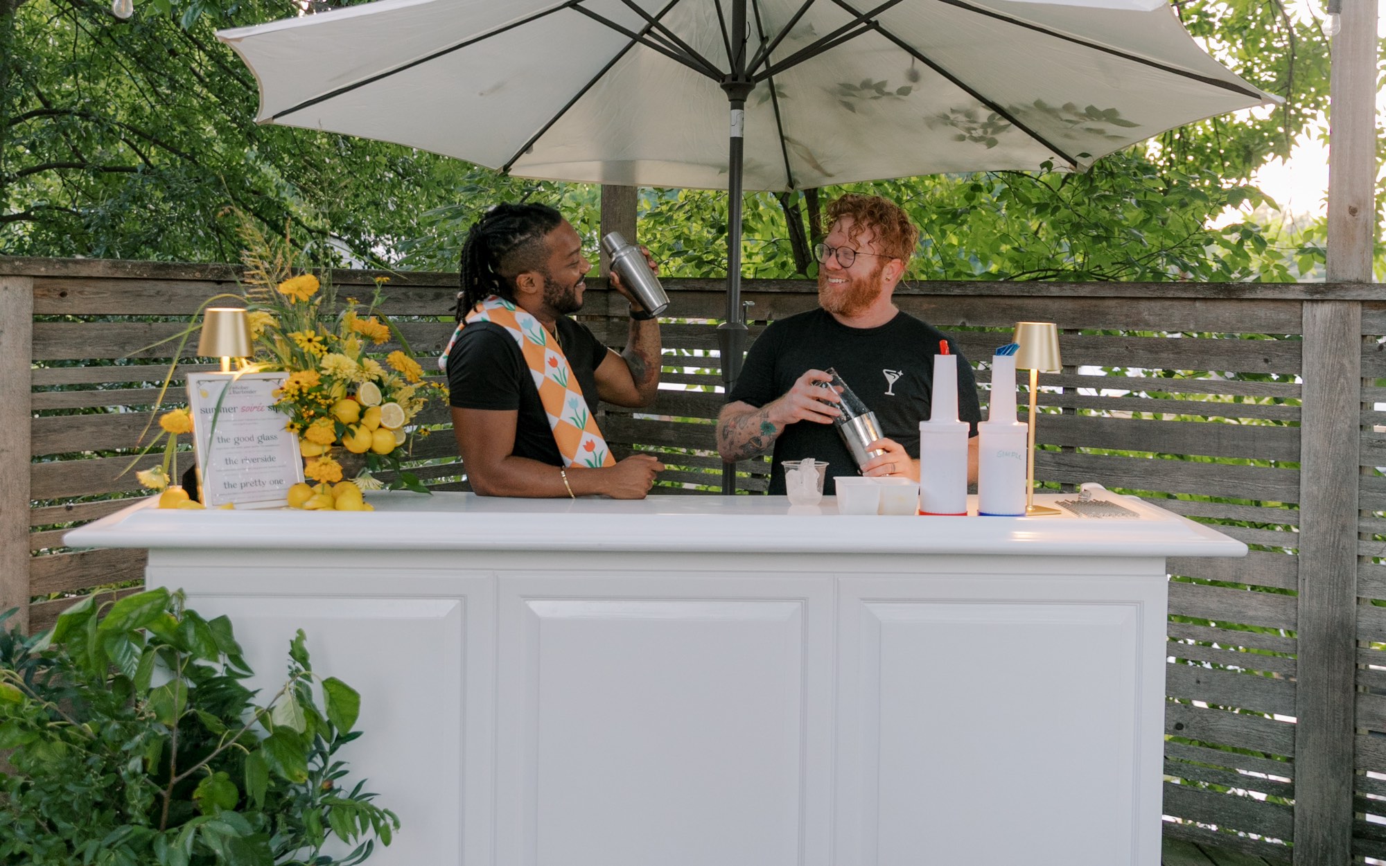 Two bartenders shaking drinks at a white bar under a patio umbrella